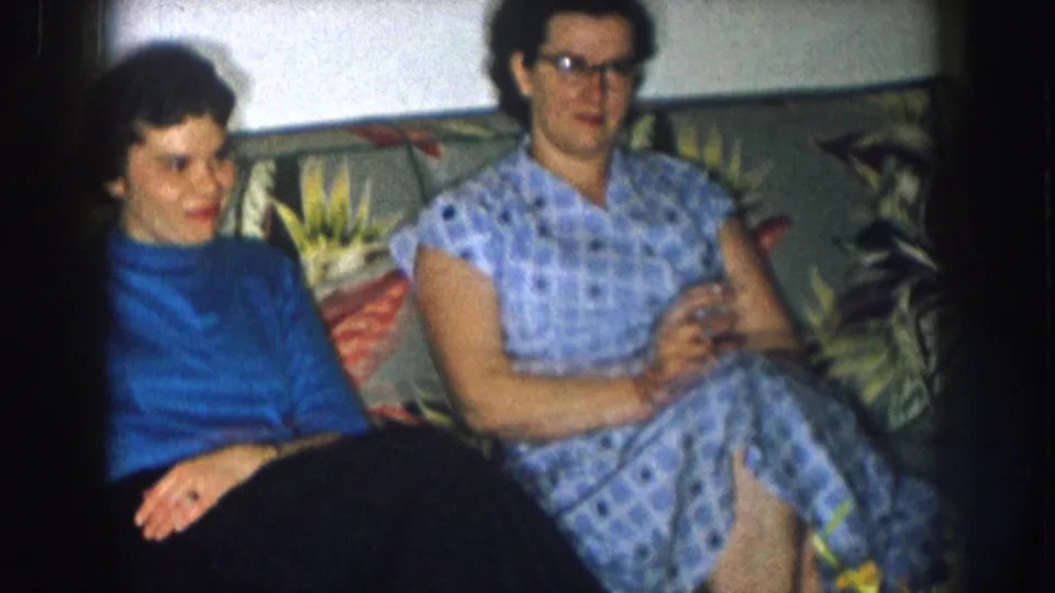 Two women on a floral couch in what appears to be a mid-1950s Nebraska living room — one in a blue turtleneck, one in a checkered dress and cat-eye glasses.