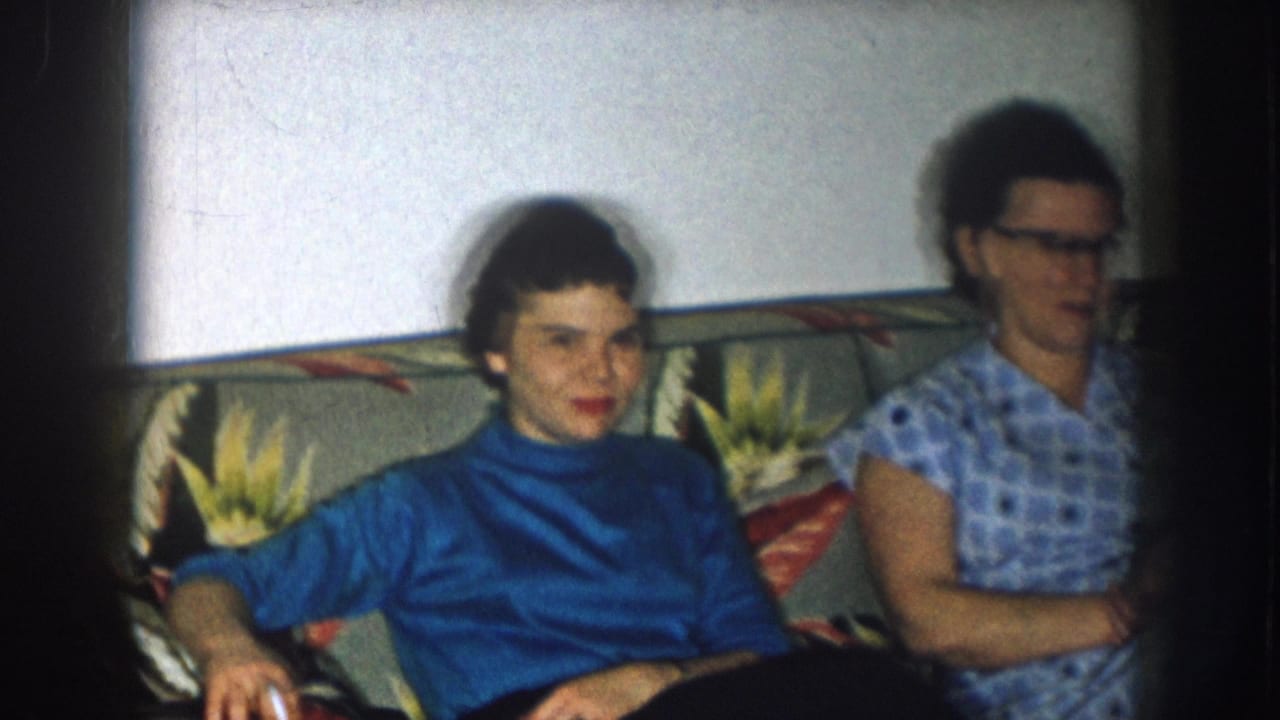 Two women on a floral-upholstered couch in what appears to be a 1950s domestic interior — the woman in a blue turtleneck faces camera directly.