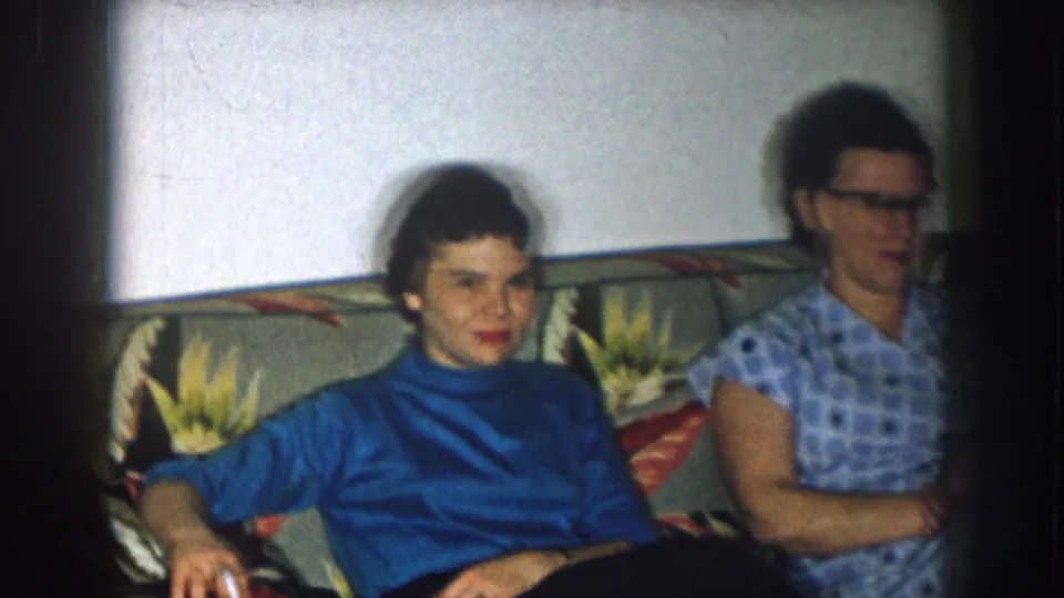 Two women on a floral-upholstered couch in what appears to be a 1950s domestic interior — the woman in a blue turtleneck faces camera directly.