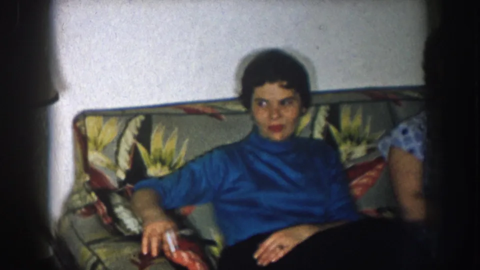 Woman in blue turtleneck seated on a floral-upholstered couch in what appears to be a 1950s American living room, with a floor lamp partially visible at right.