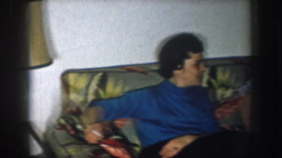 Two women in what appears to be a 1950s living room, seated on a floral couch — woman in blue turtleneck at left, woman with cat-eye glasses at right.