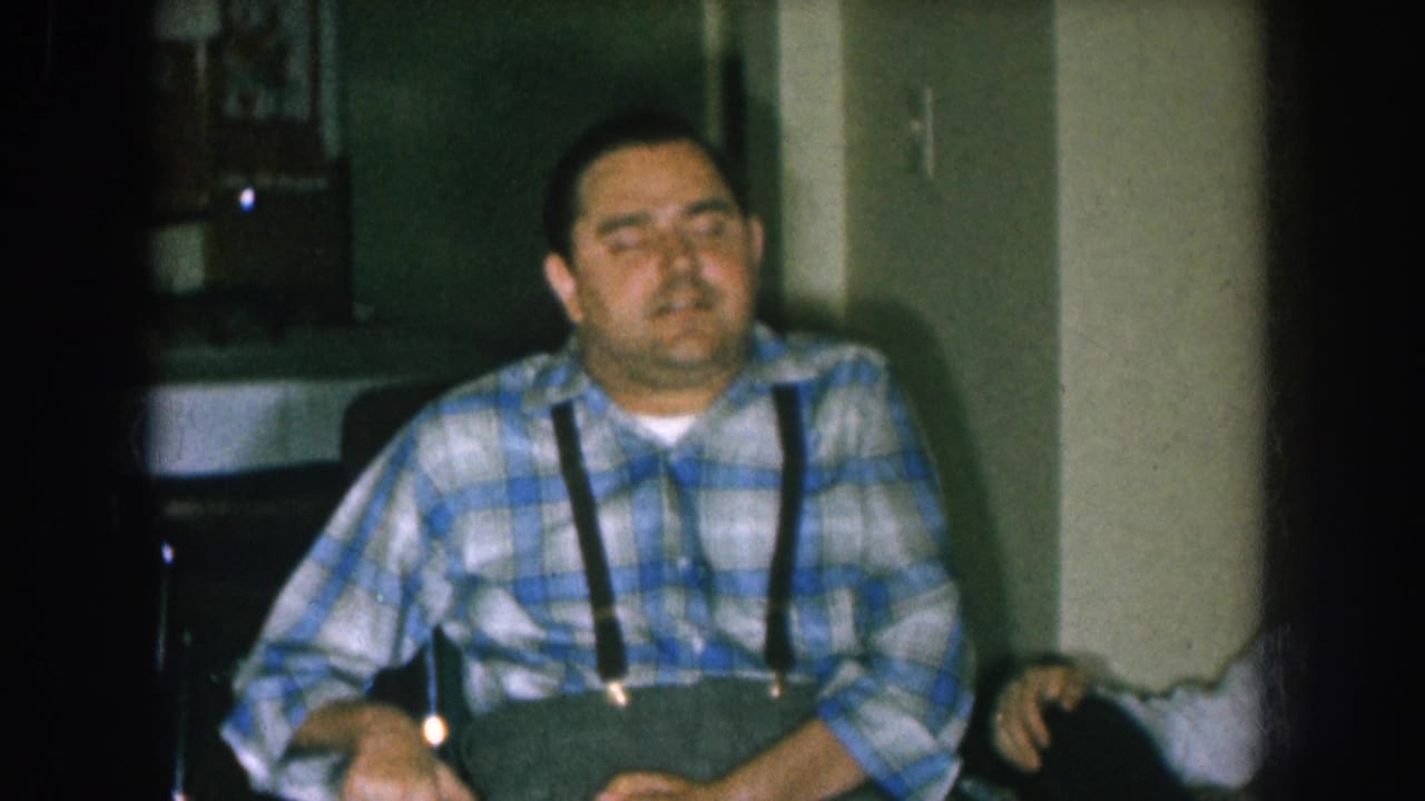 Middle-aged man in blue-and-white plaid shirt and black suspenders seated in a wheelchair in a domestic interior, Nebraska, circa 1957, shot on aged 8mm home-movie film.