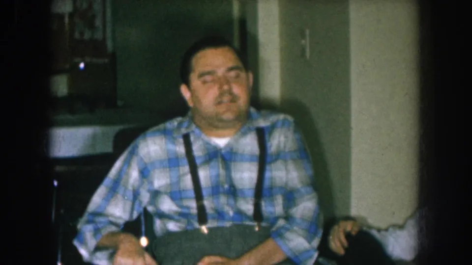 Middle-aged man in blue-and-white plaid shirt and black suspenders seated in a wheelchair in a domestic interior, Nebraska, circa 1957, shot on aged 8mm home-movie film.