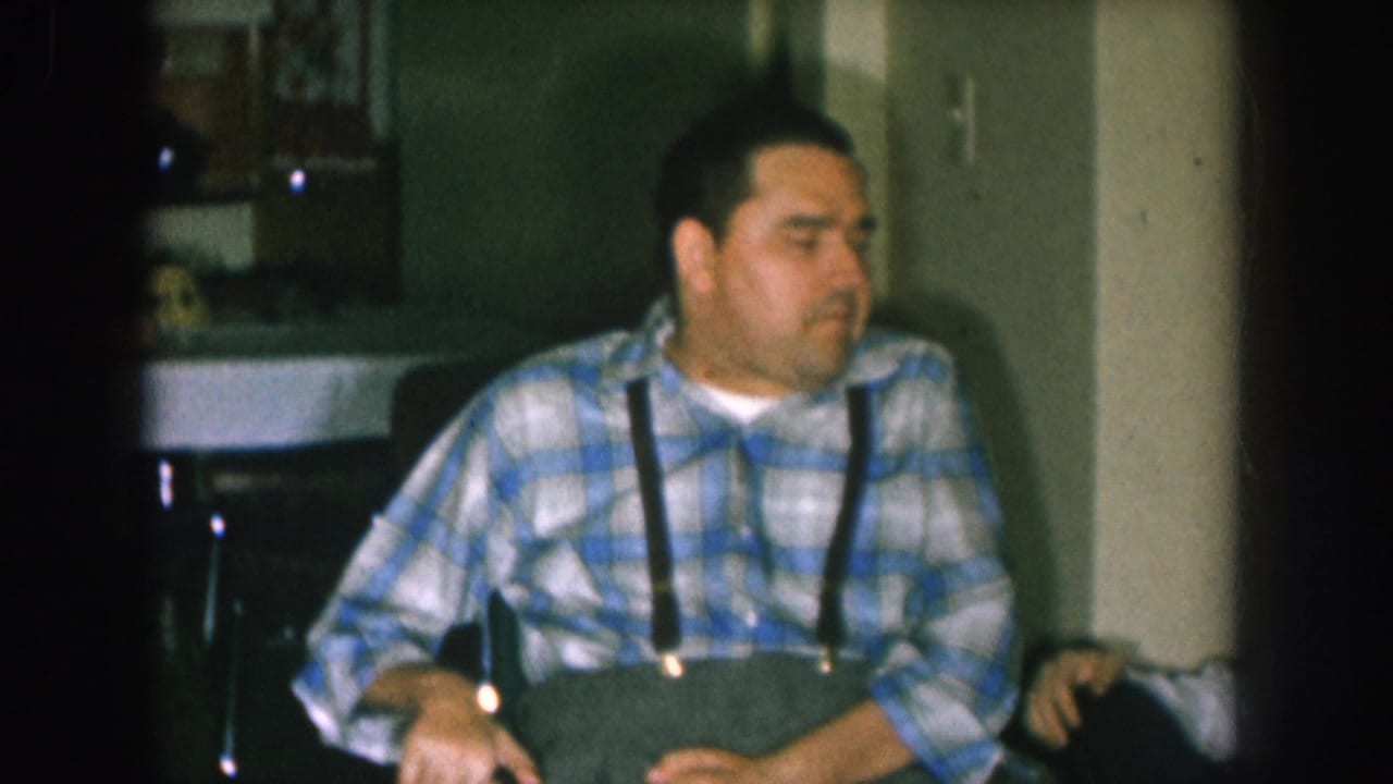 Man in plaid shirt and suspenders in wheelchair, relaxed expression, domestic background with shelving visible at upper right.