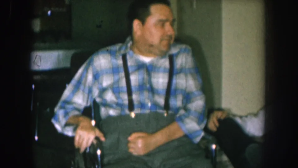 Middle-aged man in blue-and-white plaid shirt seated in a wheelchair with hands resting in lap, domestic interior, circa 1957 Nebraska, 8mm home movie.