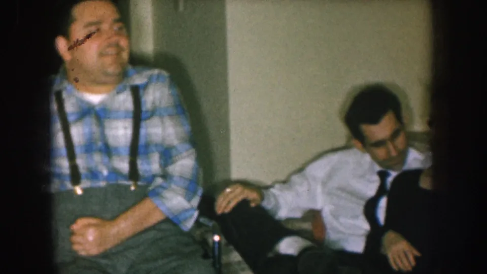 Man in blue plaid shirt and suspenders in wheelchair smiling; second individual in white shirt and dark tie partially visible at frame edge, domestic interior, circa 1957 Nebraska.