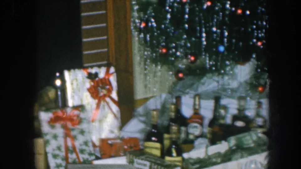 1957 Nebraska home movie: pan revealing wrapped gifts, red ribbon bows, spirit bottles, and the base of a silver-tinsel Christmas tree strung with colored fairy lights.