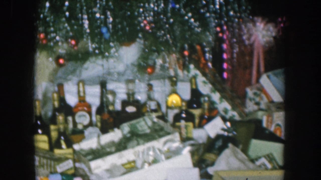 1957 Nebraska home movie: pan across spirit bottles and wrapped gifts under a silver-tinsel Christmas tree strung with red and blue fairy lights, pink ribbon bow at center.