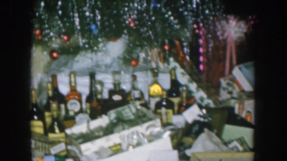 1957 Nebraska home movie: pan across spirit bottles and wrapped gifts under a silver-tinsel Christmas tree strung with red and blue fairy lights, pink ribbon bow at center.
