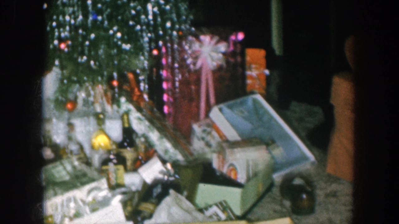 1957 Nebraska home movie: final pan position showing spirit bottles, gift boxes, and tinsel tree with fairy lights; a partial dark silhouette at the right frame edge requires human review.