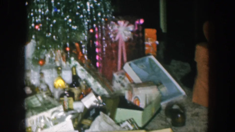 1957 Nebraska home movie: final pan position showing spirit bottles, gift boxes, and tinsel tree with fairy lights; a partial dark silhouette at the right frame edge requires human review.