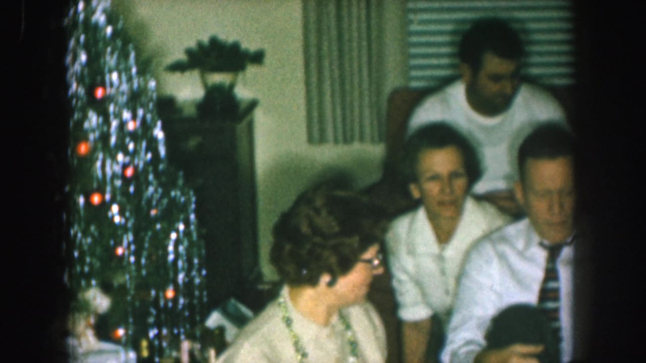 1957 Nebraska home-movie frame showing adults seated in a living room near a tinsel Christmas tree as they gather around a television set.