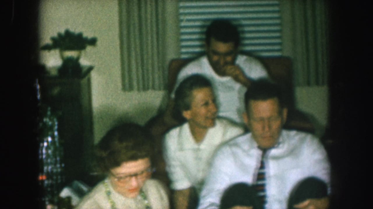 1957 Nebraska home-movie frame of adults in mid-century dress seated together in a private living room during a Christmas television gathering.