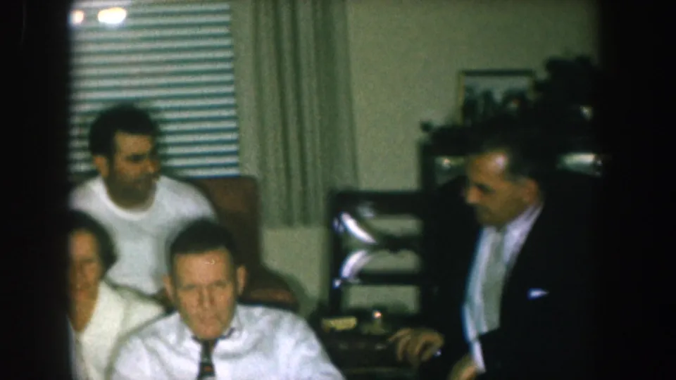 1957 Nebraska home-movie frame with a man in a dark suit entering near the television as other adults sit in a Christmas Eve living-room gathering.