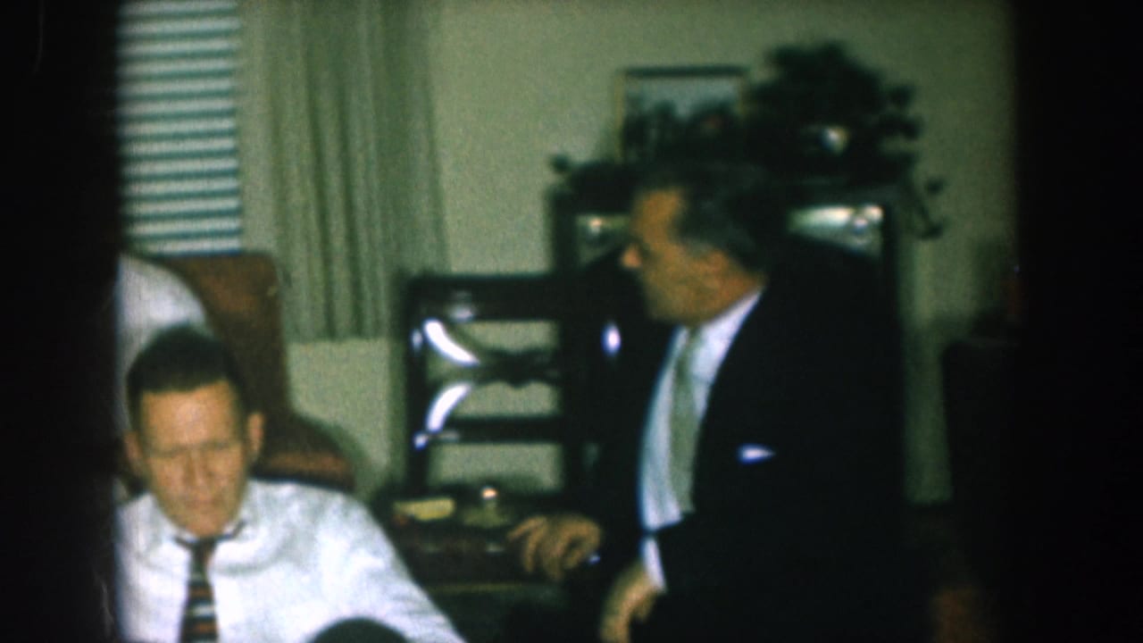 Two adult men in mid-1950s business attire converse in a domestic living room, likely Nebraska circa 1957, with ladder-back chairs and curtains visible behind them.