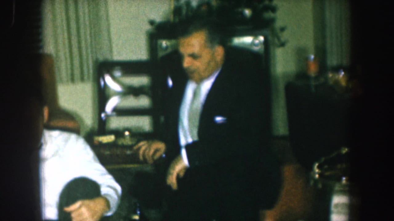 Man in dark suit stands while a figure crouches at floor level; a seated man and wooden chairs are visible in the background.