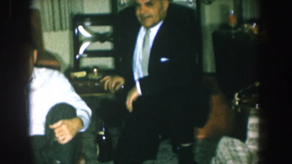 A man in a mid-1950s dark suit stands in a domestic living room as a child crouches at his feet, with another man in a white shirt partially visible at the frame edge.