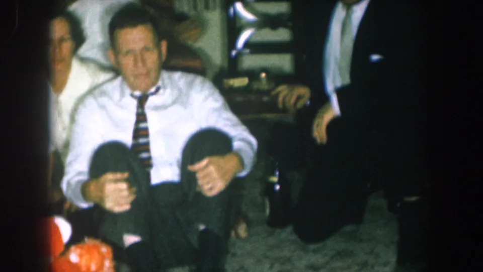 Mid-1950s domestic scene: a man in a white shirt and striped tie sits with two children at his feet, a red-orange object on the floor, a woman behind him, in what appears to be a Nebraska living room.