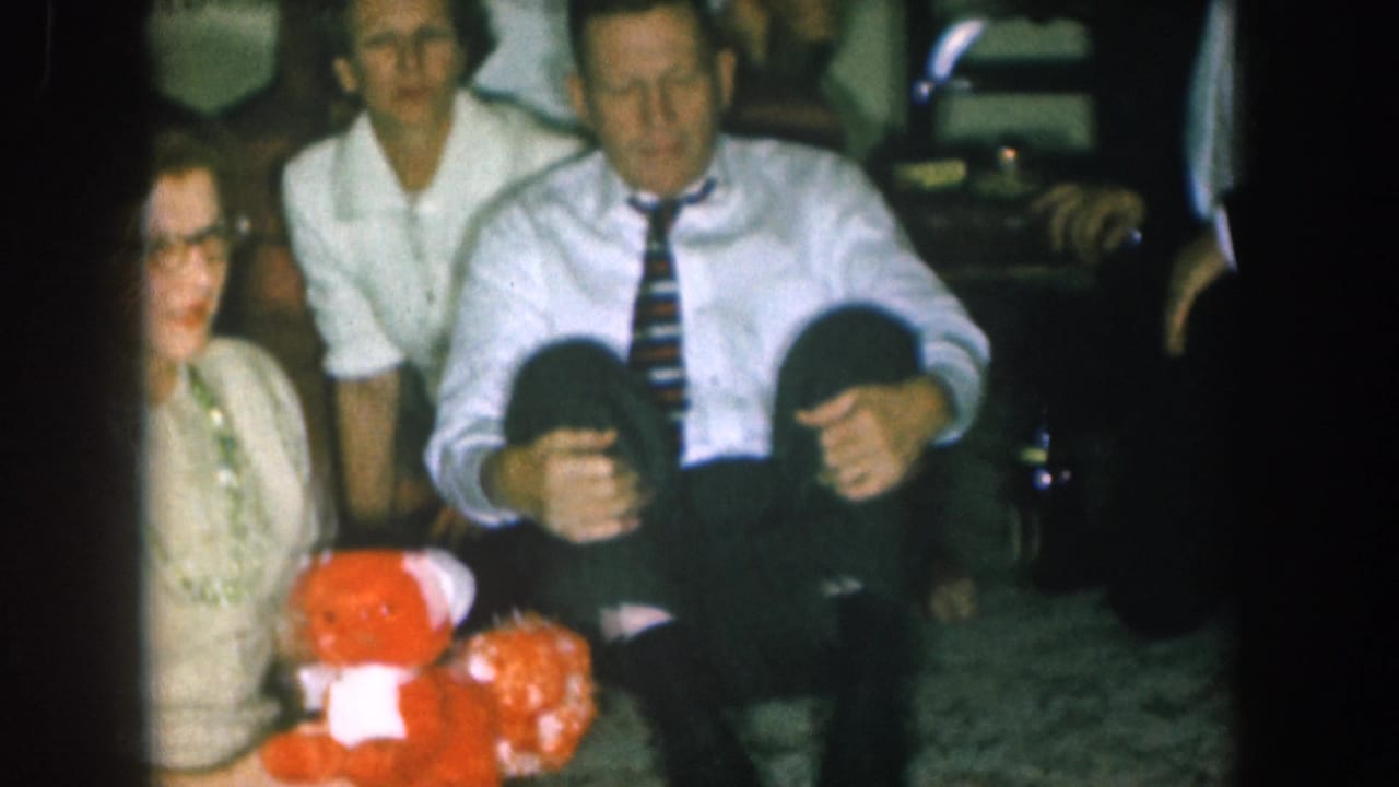 8mm home movie frame circa 1957, Nebraska: adults at a Christmas gathering with a red stuffed teddy bear; a small child appears partially visible between them.