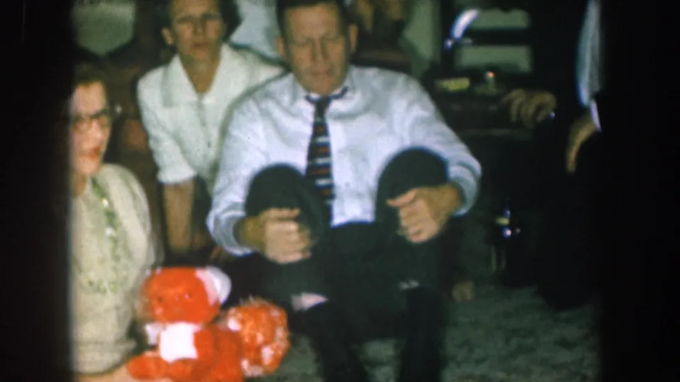 8mm home movie frame circa 1957, Nebraska: adults at a Christmas gathering with a red stuffed teddy bear; a small child appears partially visible between them.