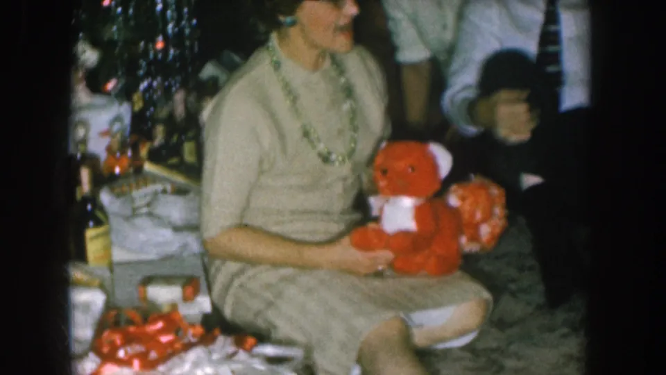 8mm home movie frame circa 1957, Nebraska: woman smiling while holding a red stuffed teddy bear gift; Christmas tree, wrapped presents, and a child partially visible nearby.