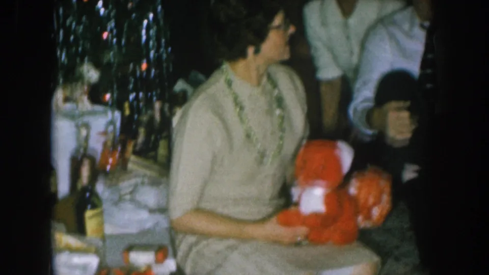 8mm home movie frame circa 1957, Nebraska: woman in tan dress holds a red stuffed teddy bear at a Christmas party; lit Christmas tree and bottles visible in background.