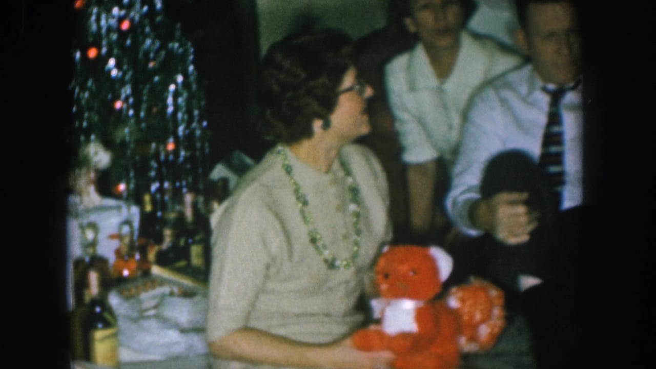 1950s Nebraska home-movie frame: woman in glasses holds a red stuffed toy bear beside a lit Christmas tree, man in striped tie visible behind her.