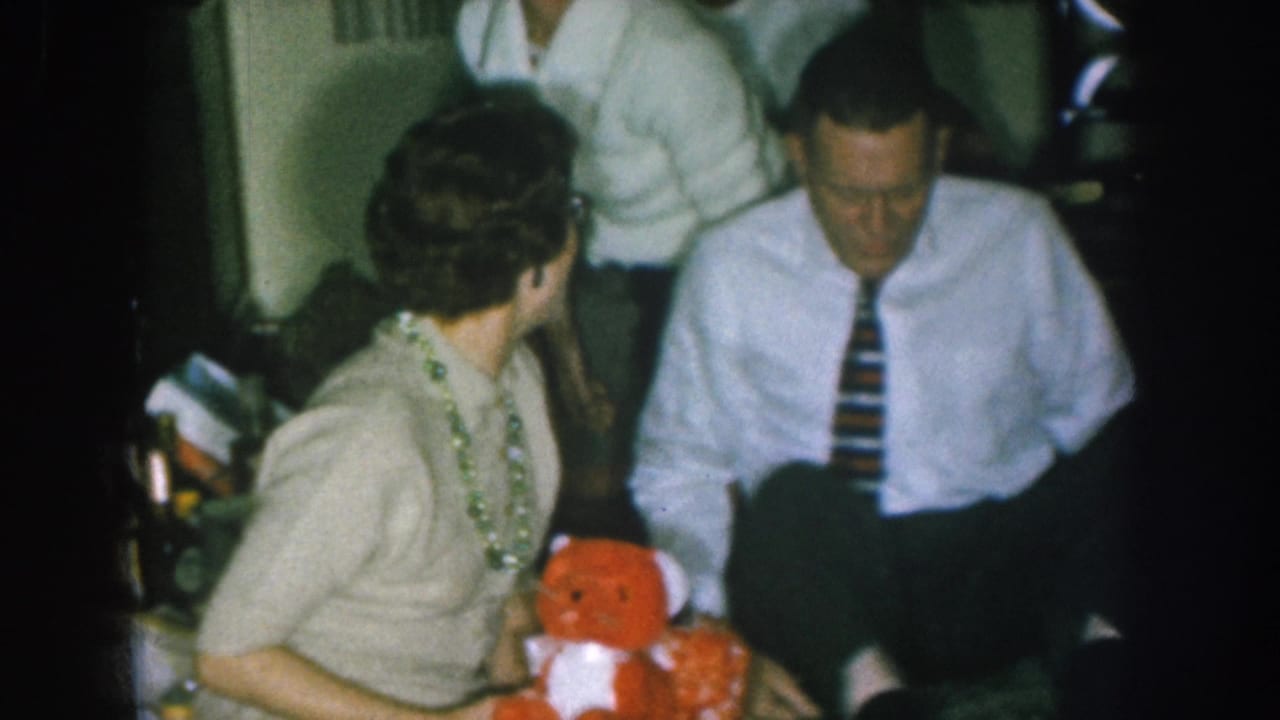 Woman in glasses and green beaded necklace holds red stuffed bear in profile; man in white shirt and striped tie seated to her right.