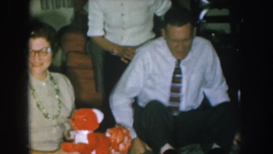 1950s Nebraska home-movie frame: woman with glasses and green necklace smiles beside a man in a striped tie leaning forward, red stuffed toy bear visible in foreground.