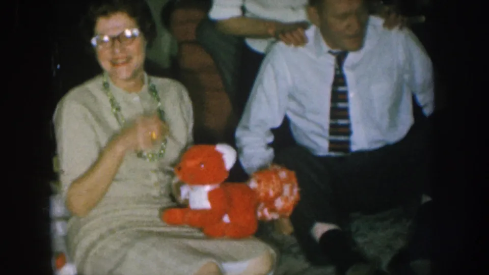 1950s Nebraska home-movie frame: woman in glasses holds red stuffed toy bear and smiles toward the camera, man in striped tie beside her.