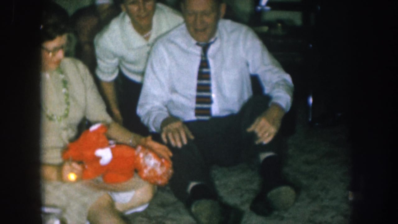 1957 Nebraska home movie: woman holds a large red stuffed toy while a man in a striped tie sits beside her, likely at a Christmas family gathering.