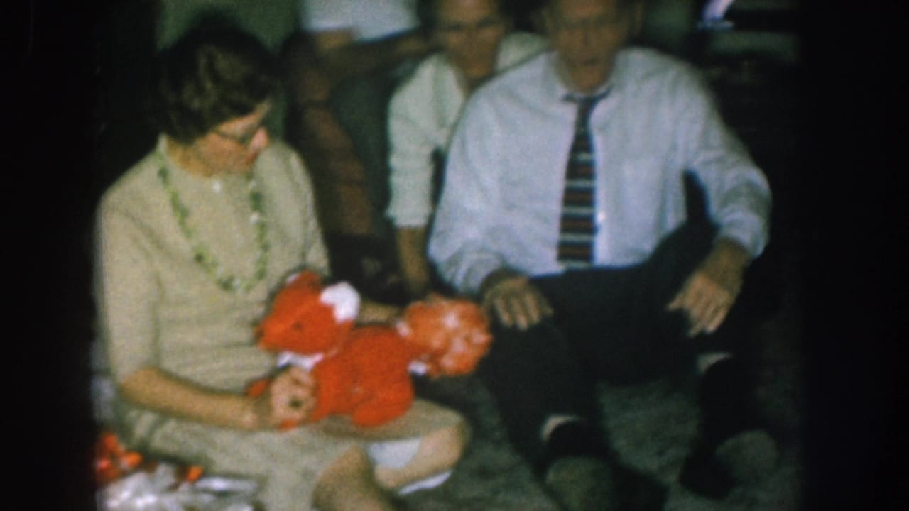 Woman with bead necklace cradles a large red stuffed animal; man in white shirt beside her; lit Christmas tree with colored lights visible in background.