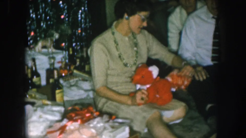 1957 Nebraska home movie: wider view of woman in glasses holding a red stuffed toy near a decorated Christmas tree; table with bottles at left; man in striped tie behind her.