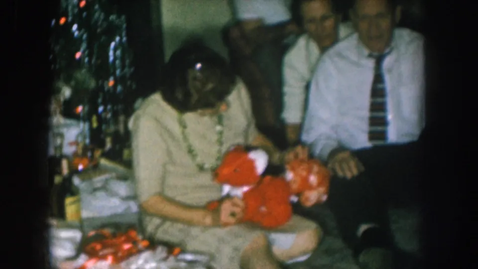 1957 Nebraska home movie: woman cradles a large red stuffed toy in a domestic interior; Christmas tree with colored lights at right; man in striped tie seated beside her.