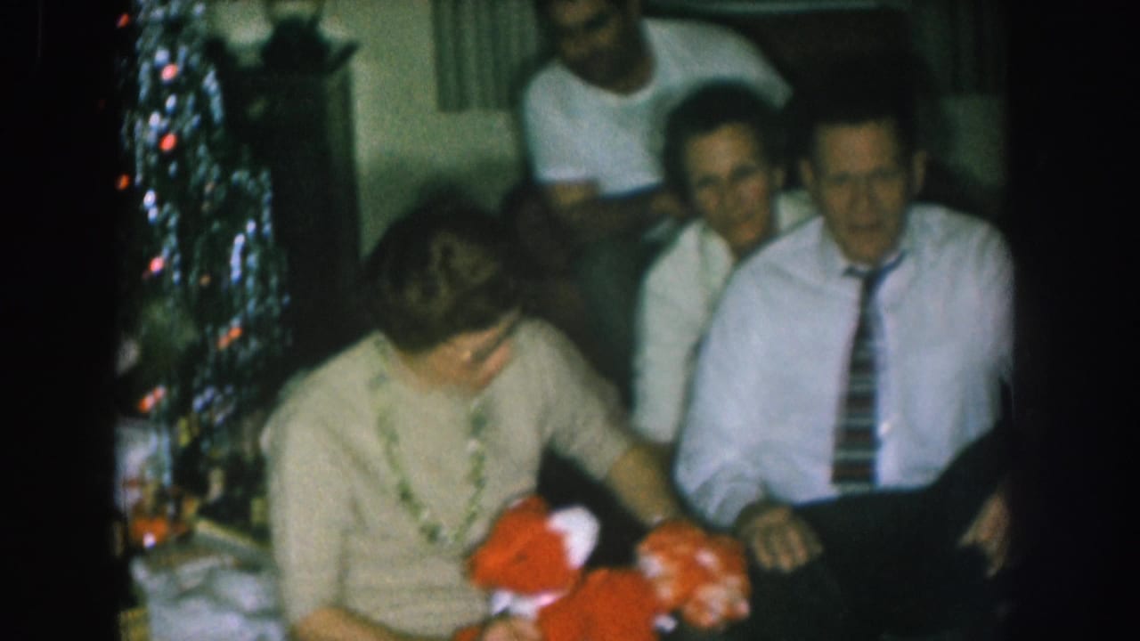 Woman in light cardigan holds a red stuffed toy while a man in a striped tie and another man in a white t-shirt watch nearby.