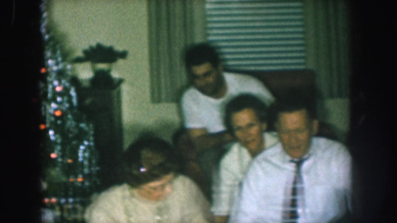 1957 Nebraska home movie still: woman with glasses focuses downward at a Christmas gathering as two male relatives look on; tree lights and window blinds visible.