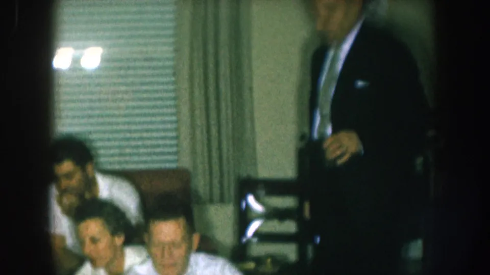 1957 Nebraska home movie still: man in dark suit stands in the living room while two seated guests appear at lower left; window blinds and chairs in background.