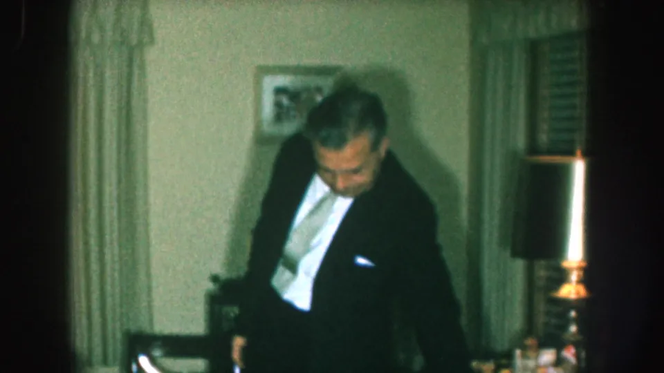 1957 Nebraska home movie still: man in dark suit with light tie looks down in a home interior; lit candle on candlestick, draped curtains, and framed wall art visible.