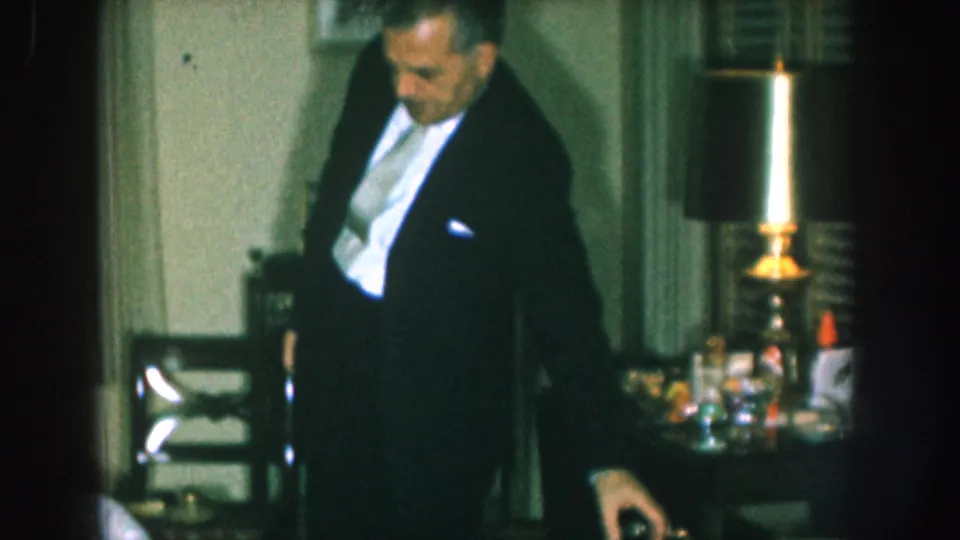 Man in dark formal jacket standing at a party table with glassware and a lit brass candlestick, 8mm home movie, likely Nebraska, 1957.