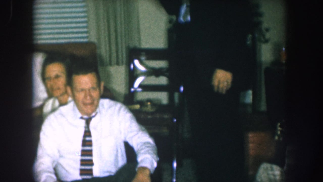 Man in white dress shirt and bold striped tie seated and smiling toward camera; second person partially visible behind him.