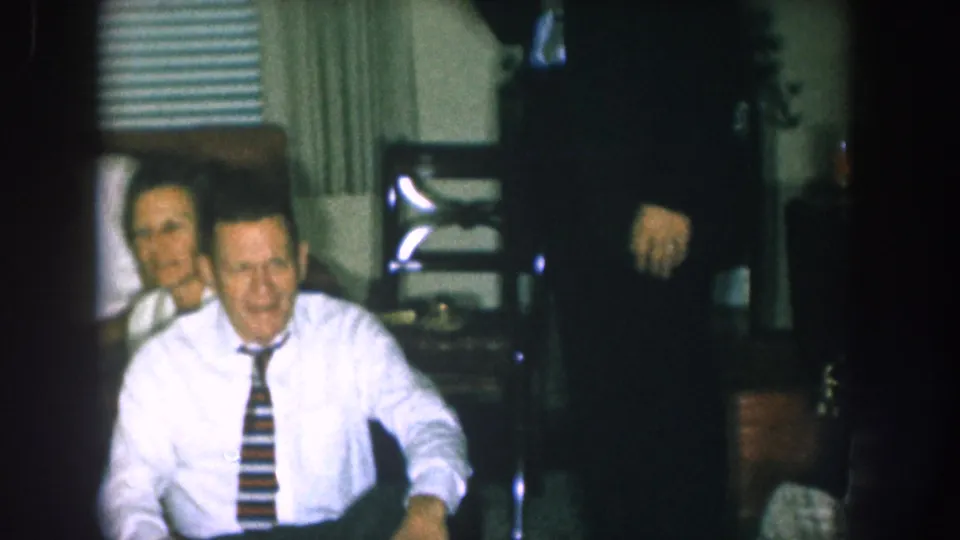 Seated man in white dress shirt and bold striped tie smiling toward camera at an indoor holiday gathering, likely Nebraska, c. 1957.