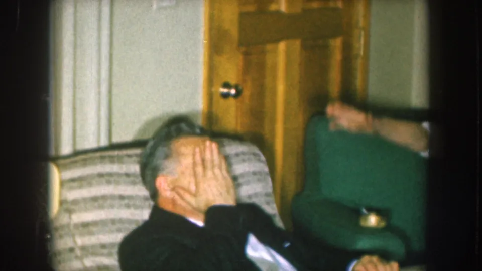 Man in dark formal wear seated on a striped sofa covering his face in apparent laughter, wooden interior door visible behind him, c. 1957.