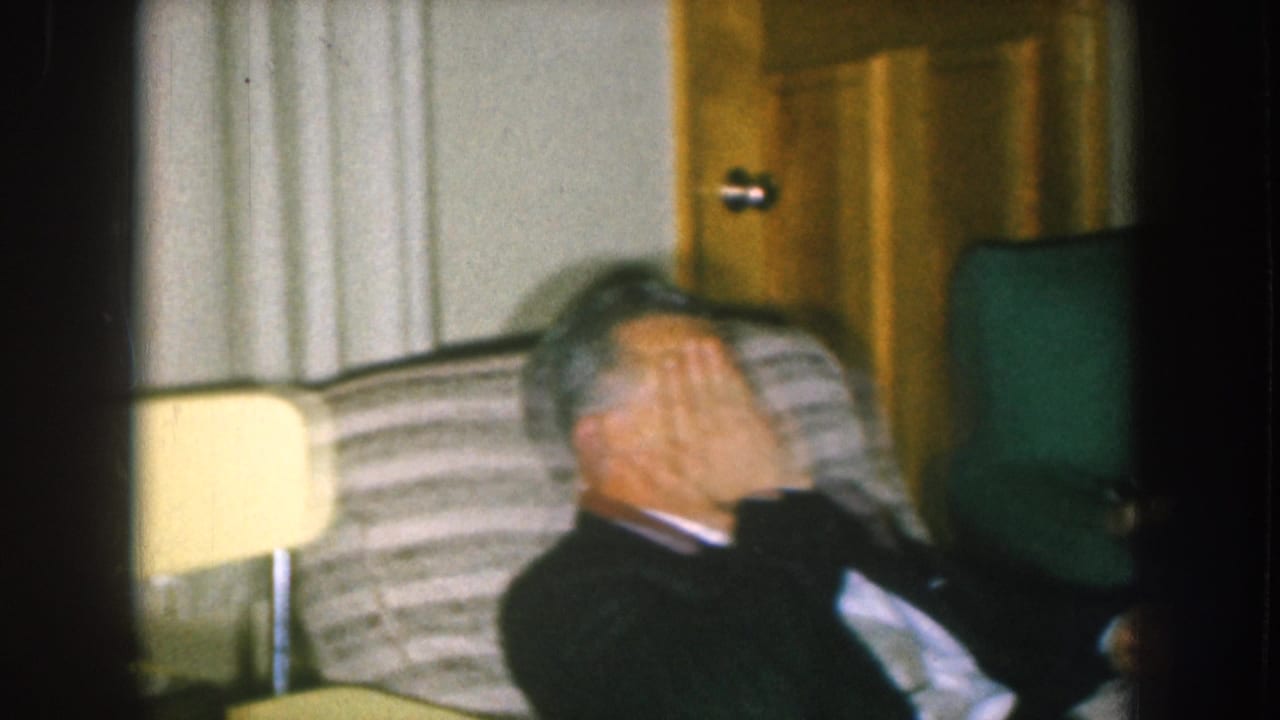 Man in dark suit covering his face mid-laugh, circa 1957, seated on a striped sofa in a mid-century Nebraska living room.