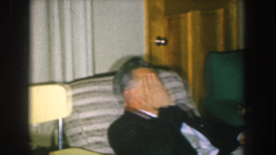 Man in dark suit covering his face mid-laugh, circa 1957, seated on a striped sofa in a mid-century Nebraska living room.