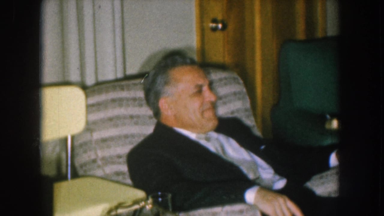 Grey-haired man in a dark suit leaning back on a striped sofa with face visible and mouth open laughing, green armchair and wood cabinet behind.