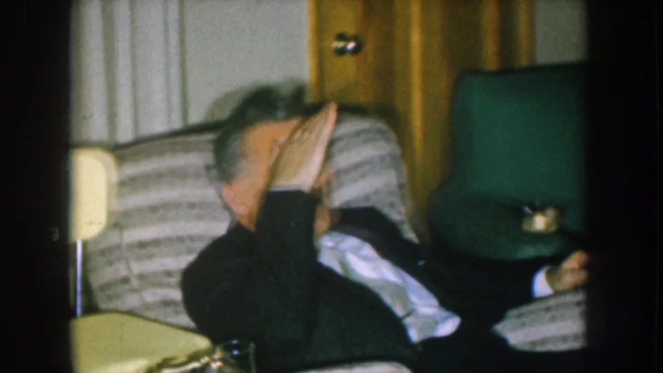 Man laughing and shielding his face with his hand, circa 1957, on a striped sofa in a mid-century Nebraska living room, cream lampshade at left.