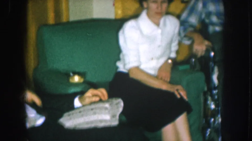 Woman in white blouse seated in a green upholstered armchair, circa 1957, in a mid-century Nebraska living room; a second person is partially visible at frame edge.