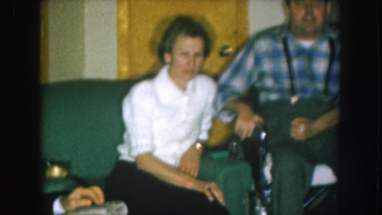 8mm color home movie circa 1957 Nebraska; woman in white shirt sits beside man in plaid shirt and suspenders in a wheelchair, with a third figure partially visible above.
