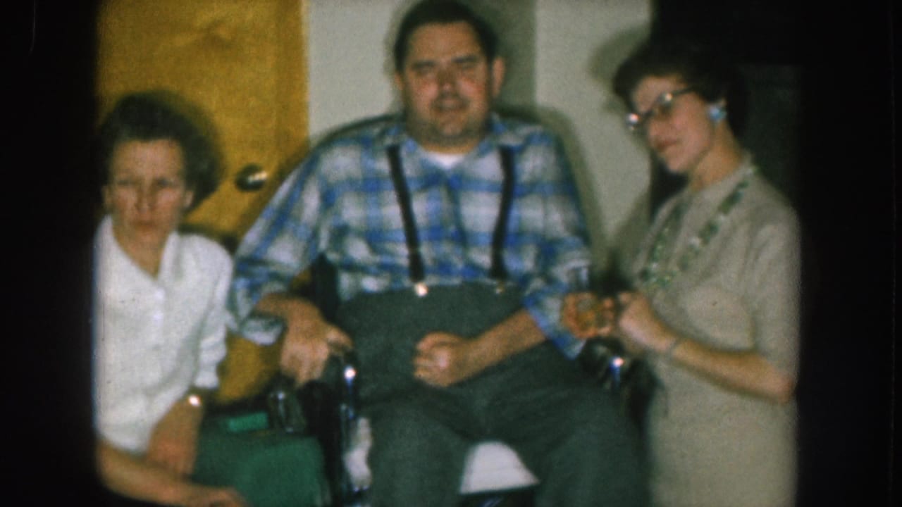 Man in plaid shirt and suspenders in a wheelchair flanked by a woman in white shirt at left and a woman wearing glasses holding a glass at right, home interior.