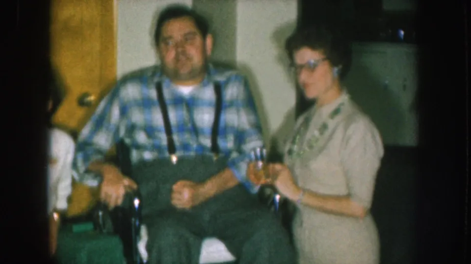 Closer framing of the same 8mm sequence circa 1957; man in wheelchair wearing blue plaid and suspenders, woman with cat-eye-style glasses in beige knitwear holding a glass.
